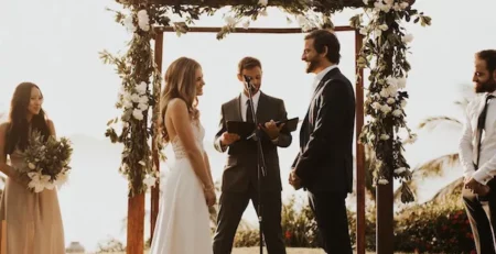 A bride and groom stand below an arbor with flowers