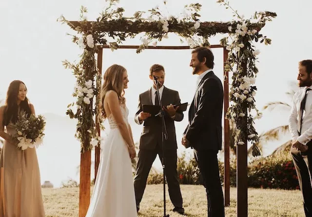 A bride and groom stand below an arbor with flowers