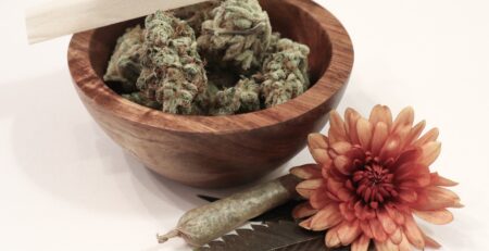 A rolling paper sits atop a wooden bowl with cannabis flower; a rolled joint and red-orange flower sit on the white surface beside the bowl.
