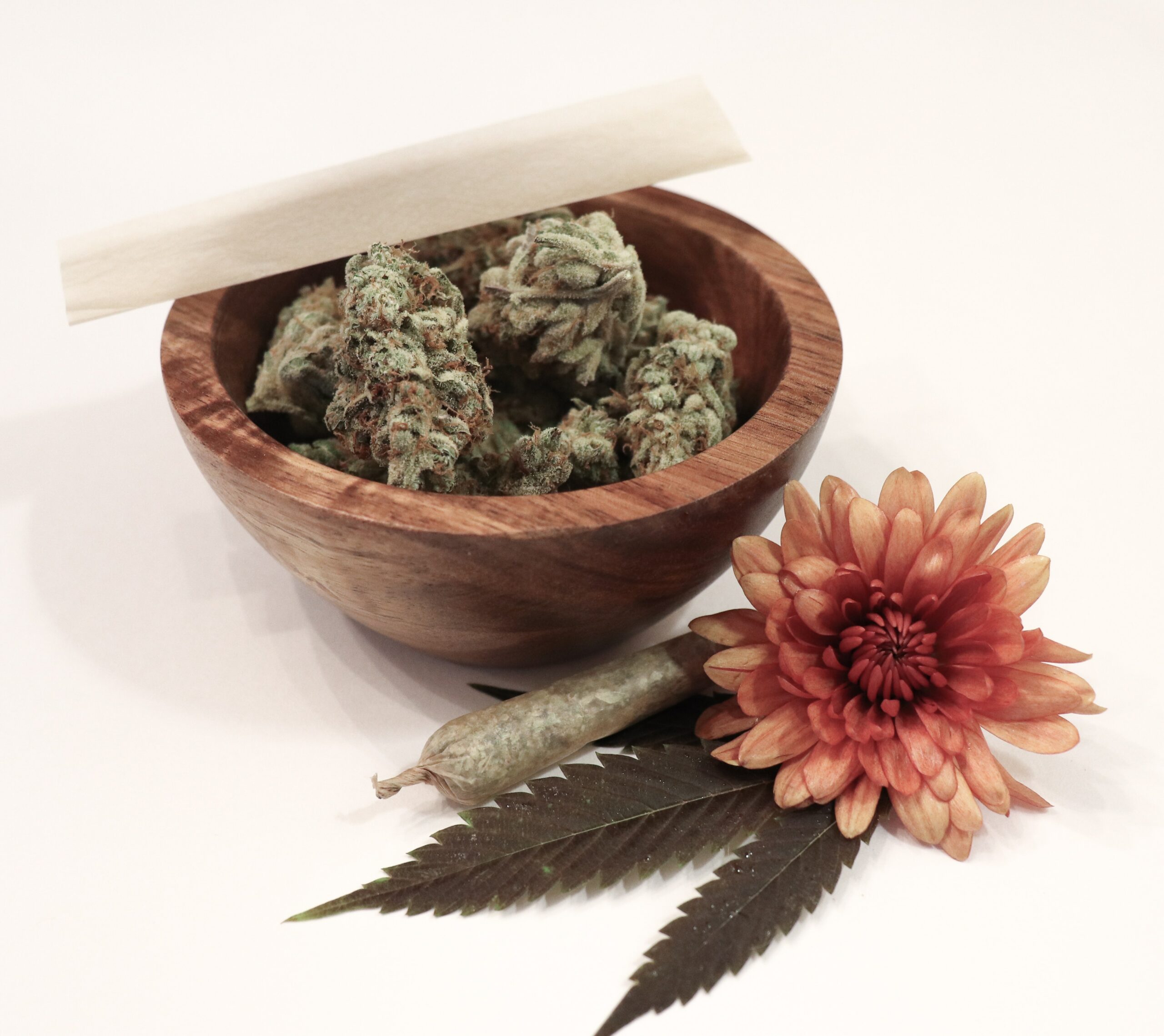 A rolling paper sits atop a wooden bowl with cannabis flower; a rolled joint and red-orange flower sit on the white surface beside the bowl.