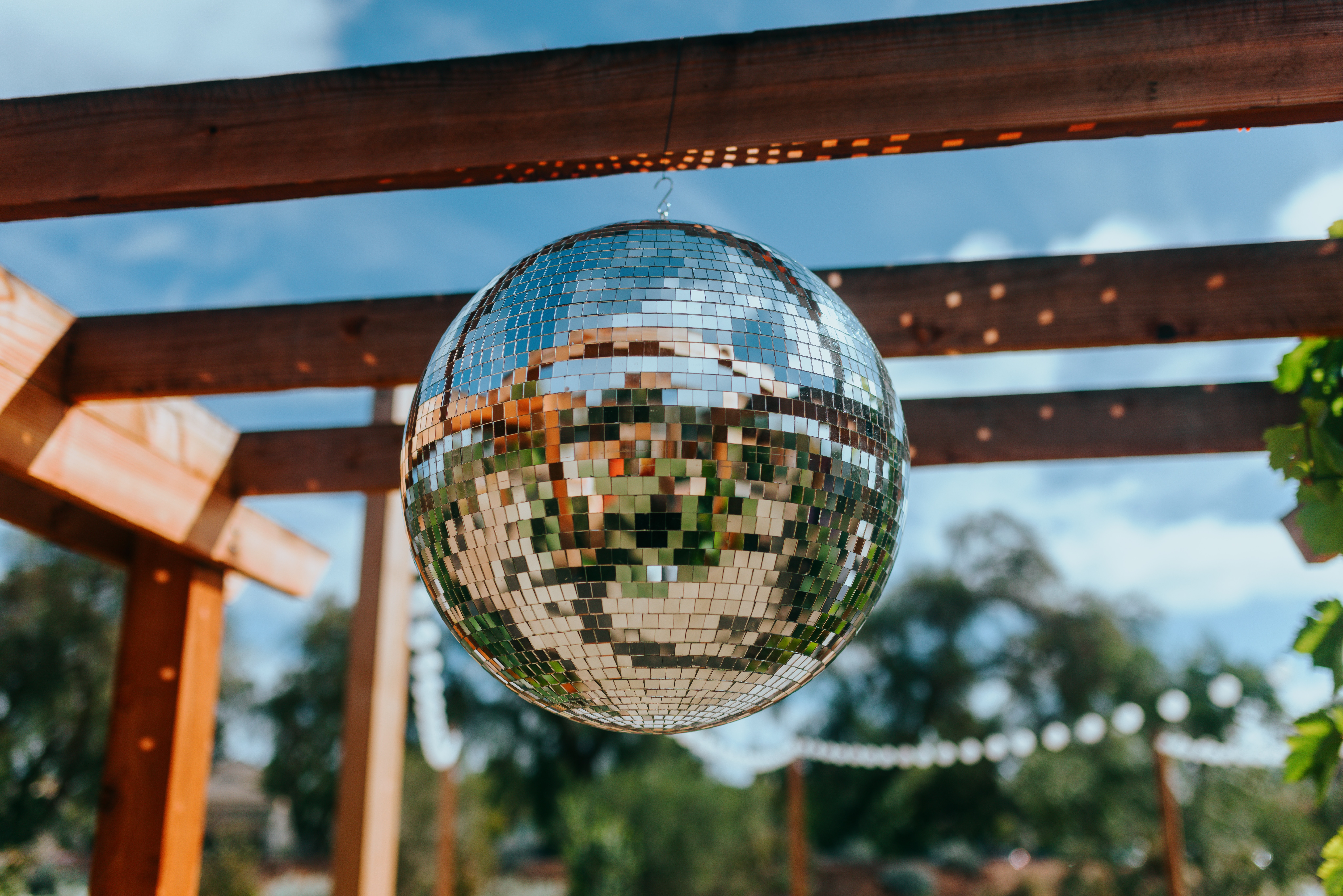 A disco ball hangs outside below a pergola, with a band of lights in the background