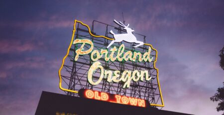 An Oregon-shaped neon sign reading 'Portland Oregon Old Town' and featuring a leaping stag perches atop a building. The sky is purple at sunset.