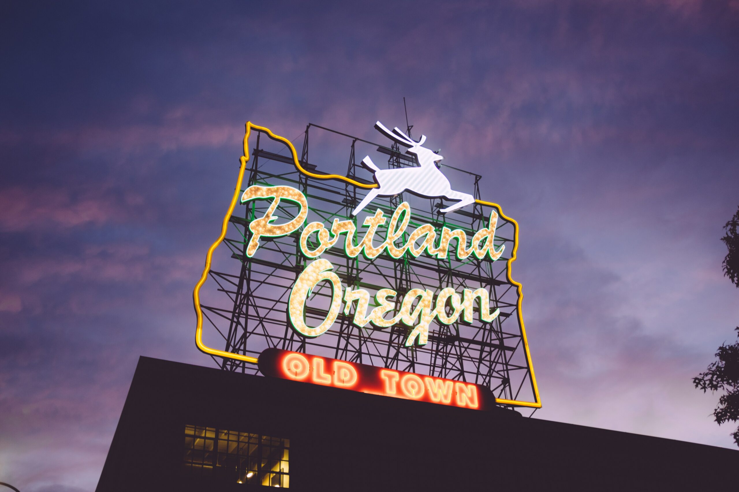 An Oregon-shaped neon sign reading 'Portland Oregon Old Town' and featuring a leaping stag perches atop a building. The sky is purple at sunset.