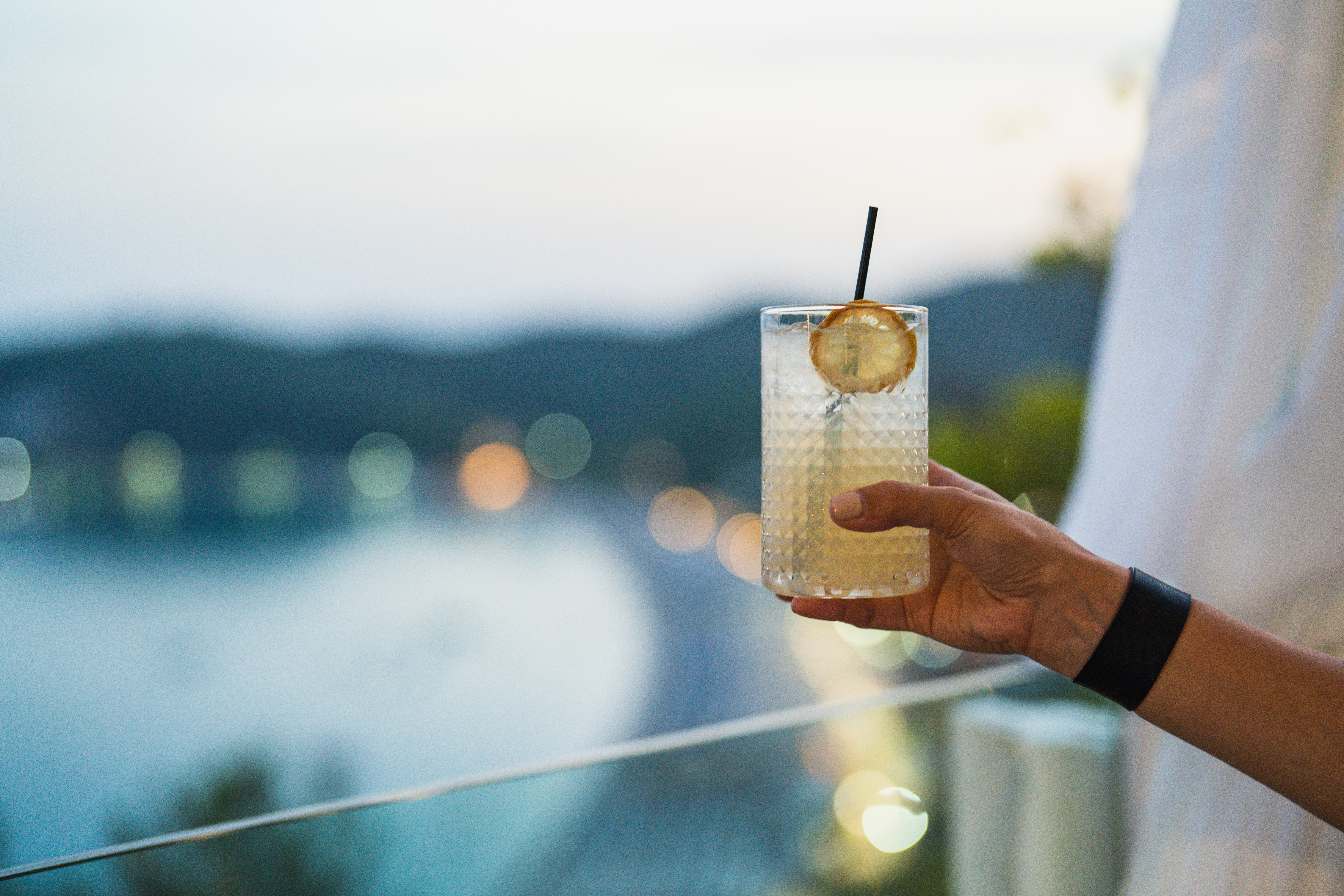 A hand with painted nails and a banded bracelet holds a cocktail in a textured glass with a straw and citrus slice against a lake and mountain backdrop.