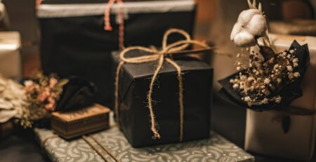 Two gifts in black wrapping paper and tied with twine sit beside cotton and other botanicals in a jar atop a counter.