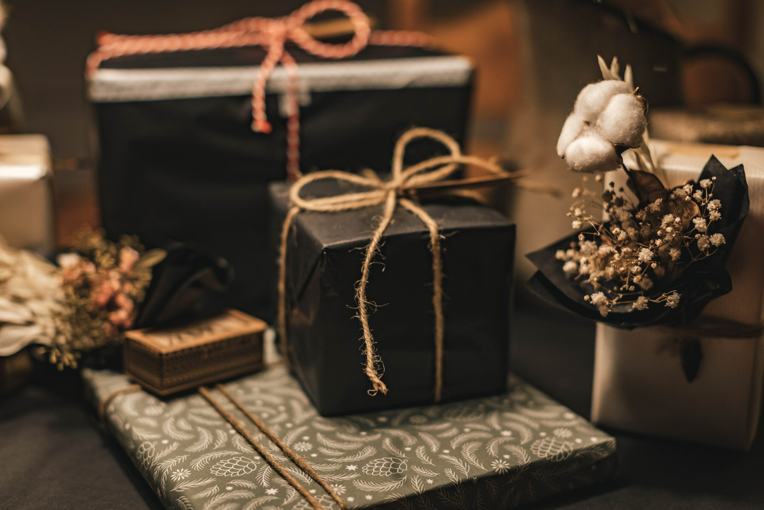 Two gifts in black wrapping paper and tied with twine sit beside cotton and other botanicals in a jar atop a counter.