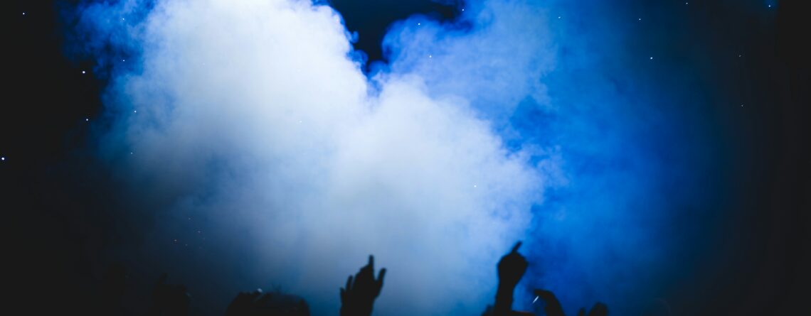 The silhouette of people raising their arms and partying at the base of a blue cloud of smoke at a stoner event.