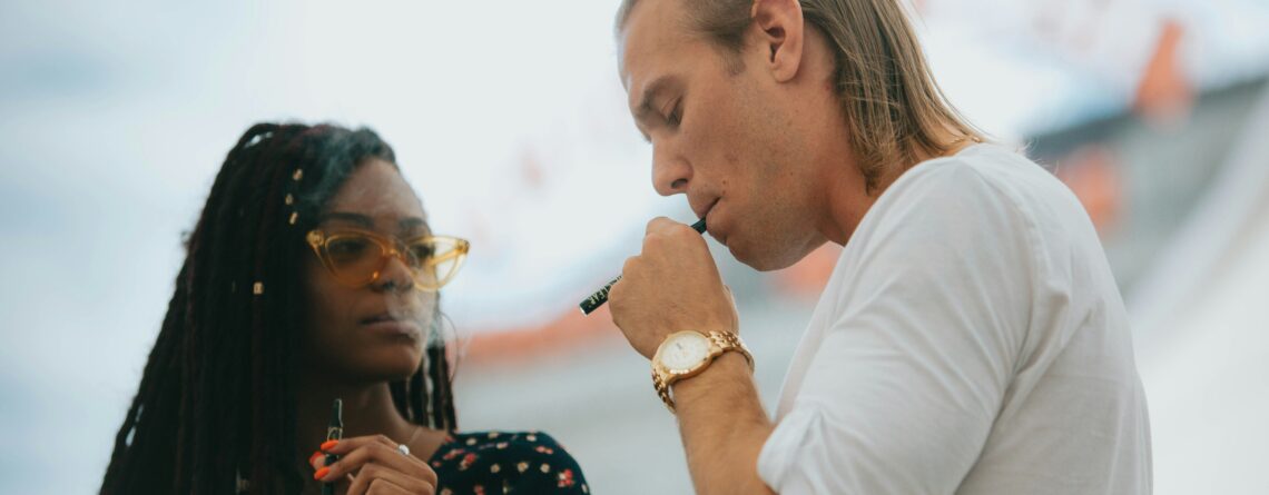 A blonde man with hair in a bun takes a rip from a cannabis vape pen; a woman with locks and sunglasses blows out a puff to the left.